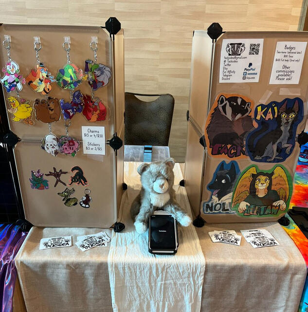 An artist's alley table setup showing charms, stickers and badges for sale.